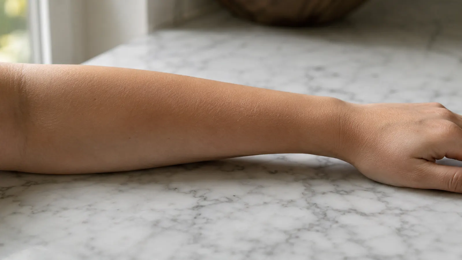Close-up of a woman's bare forearm resting on a marble bathroom counter, skin texture visible, natural morning light