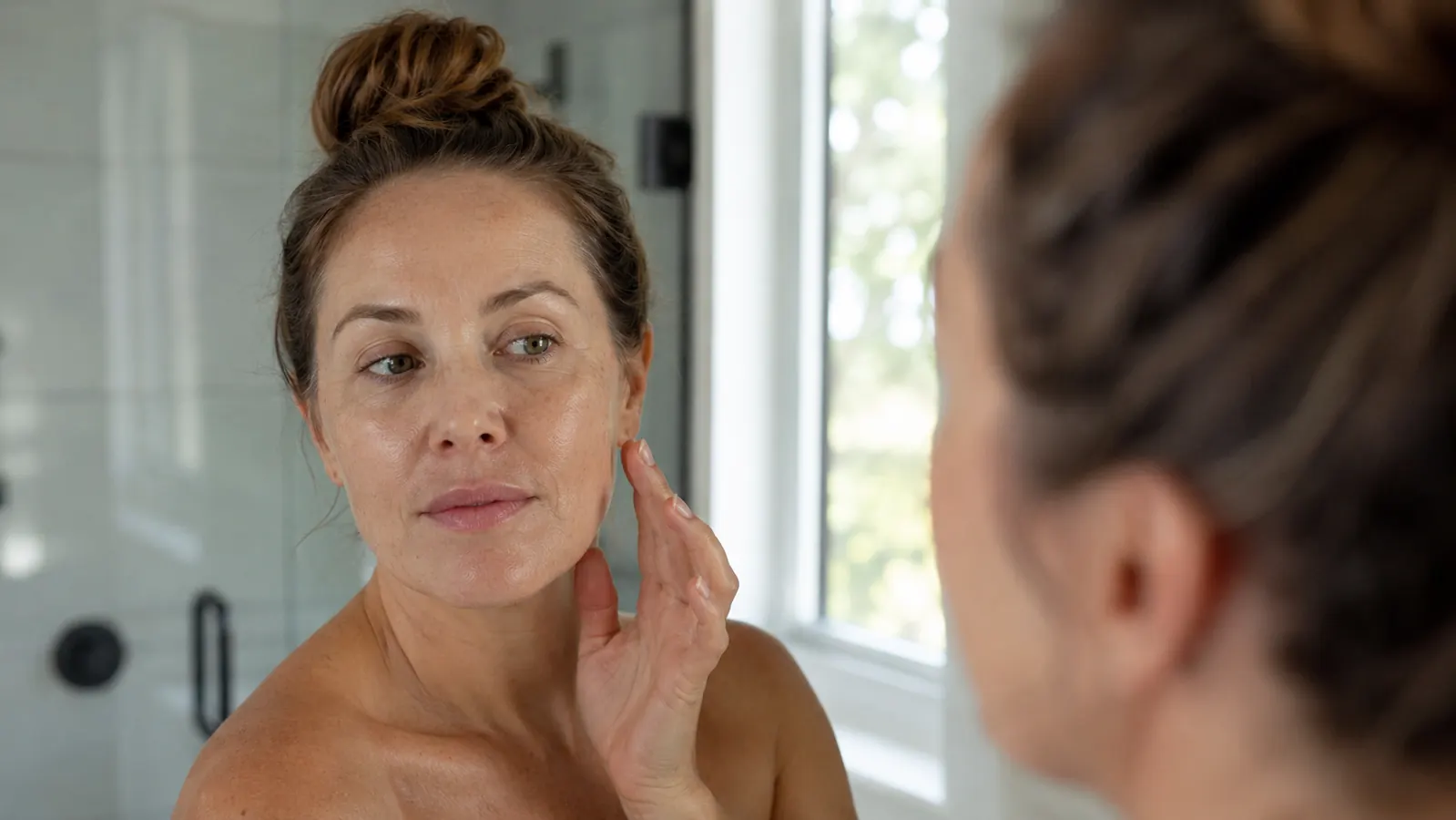 Woman with clear, calm skin looking at her face in a bathroom mirror, soft morning light