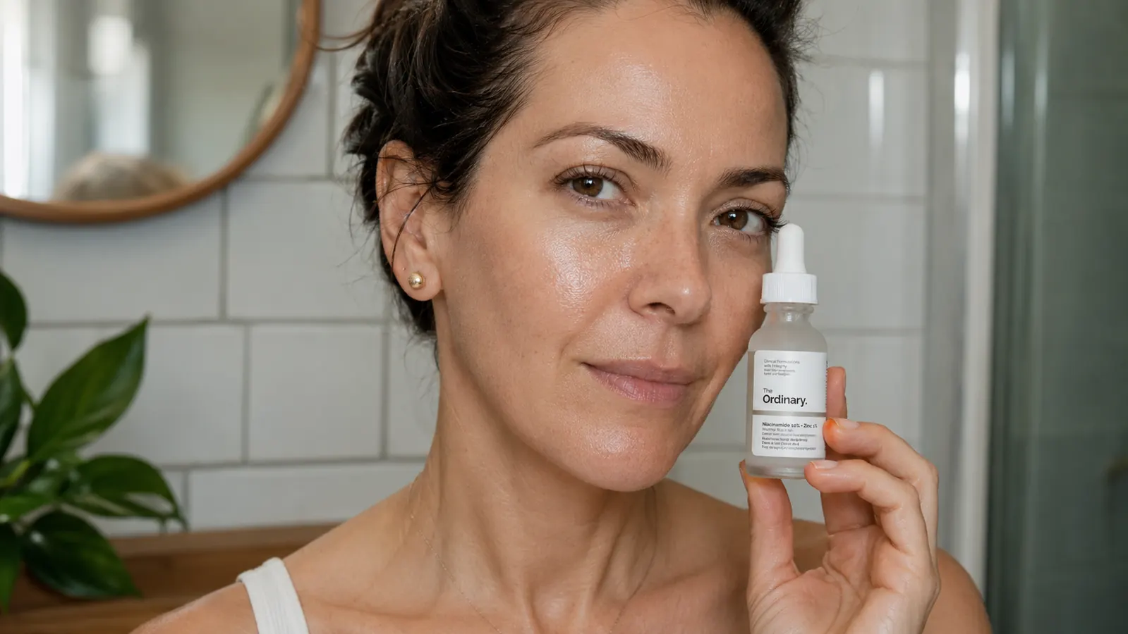 Close-up hand holding The Ordinary Niacinamide 10% + Zinc 1% serum bottle over a white bathroom counter