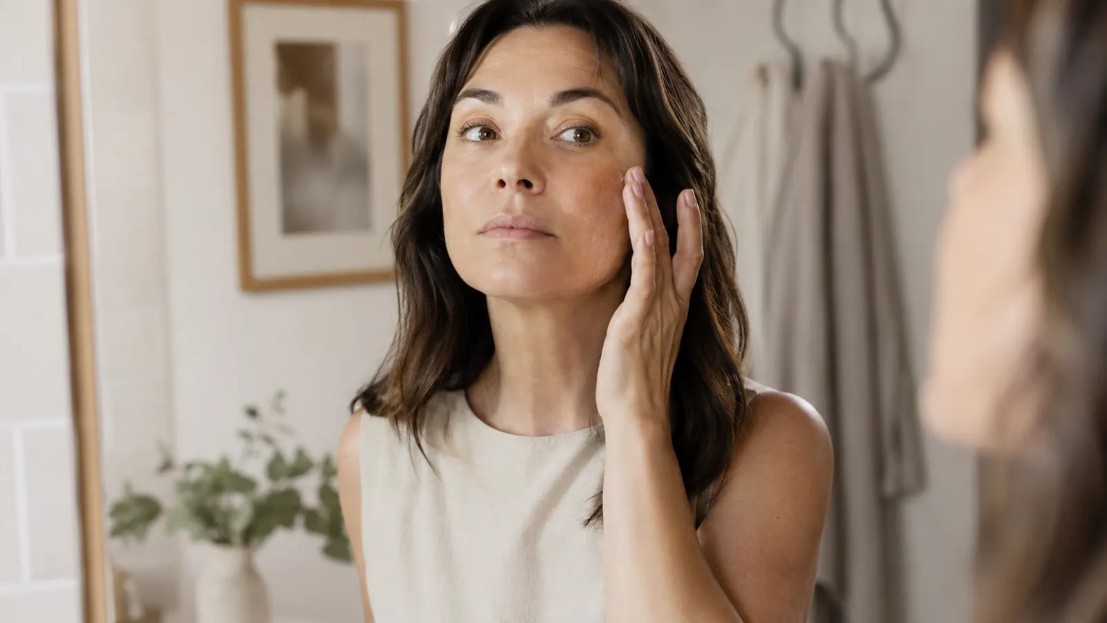 Woman in her late 30s applying serum to her face at a bathroom vanity, soft morning light coming through a frosted window